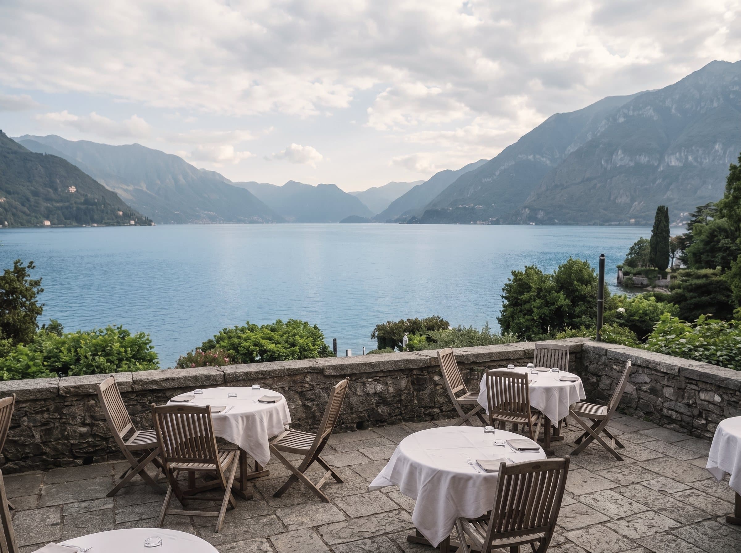 A lakeside restaurant terrace with tables laid in white linen, the lake, mountains and a wide sky beyond